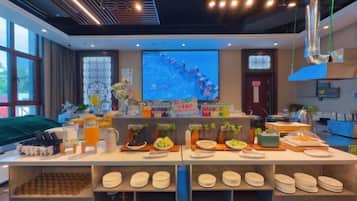 A buffet setup with various food items and beverages, a projection screen in the background, and a kitchen area.