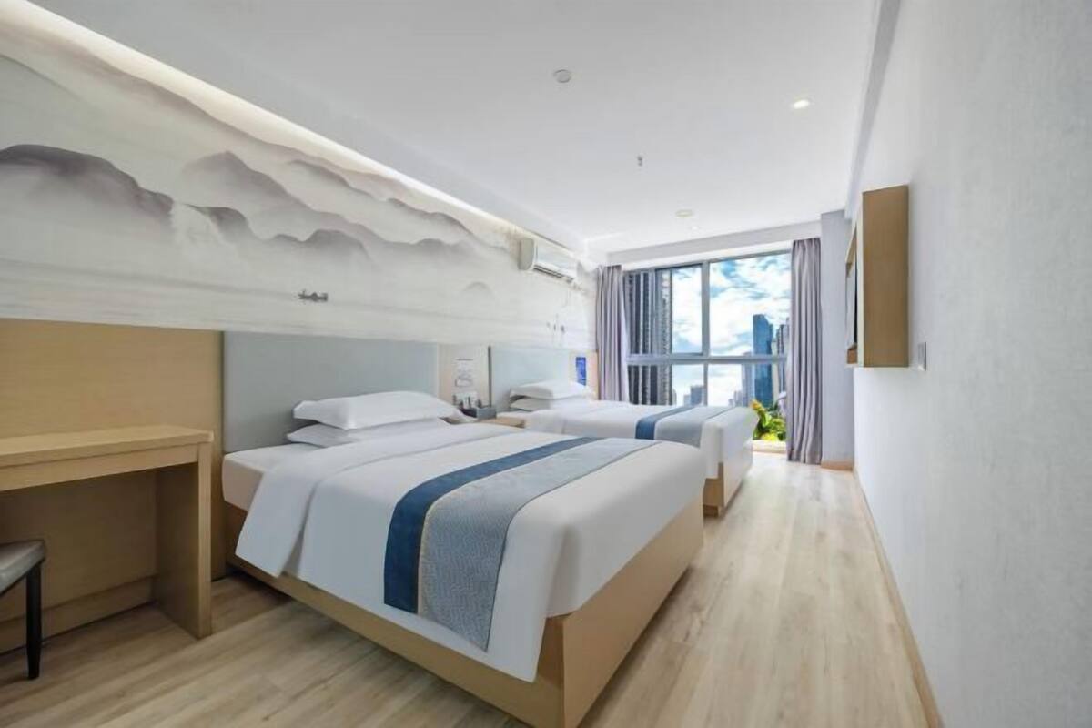 A hotel room with two beds, a desk, a window with curtains, and a wall-mounted TV.