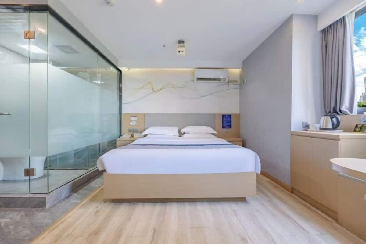 A modern hotel room with a glass shower enclosure, a bed with a wooden headboard, a bedside table, a built-in sink, and a cityscape view.