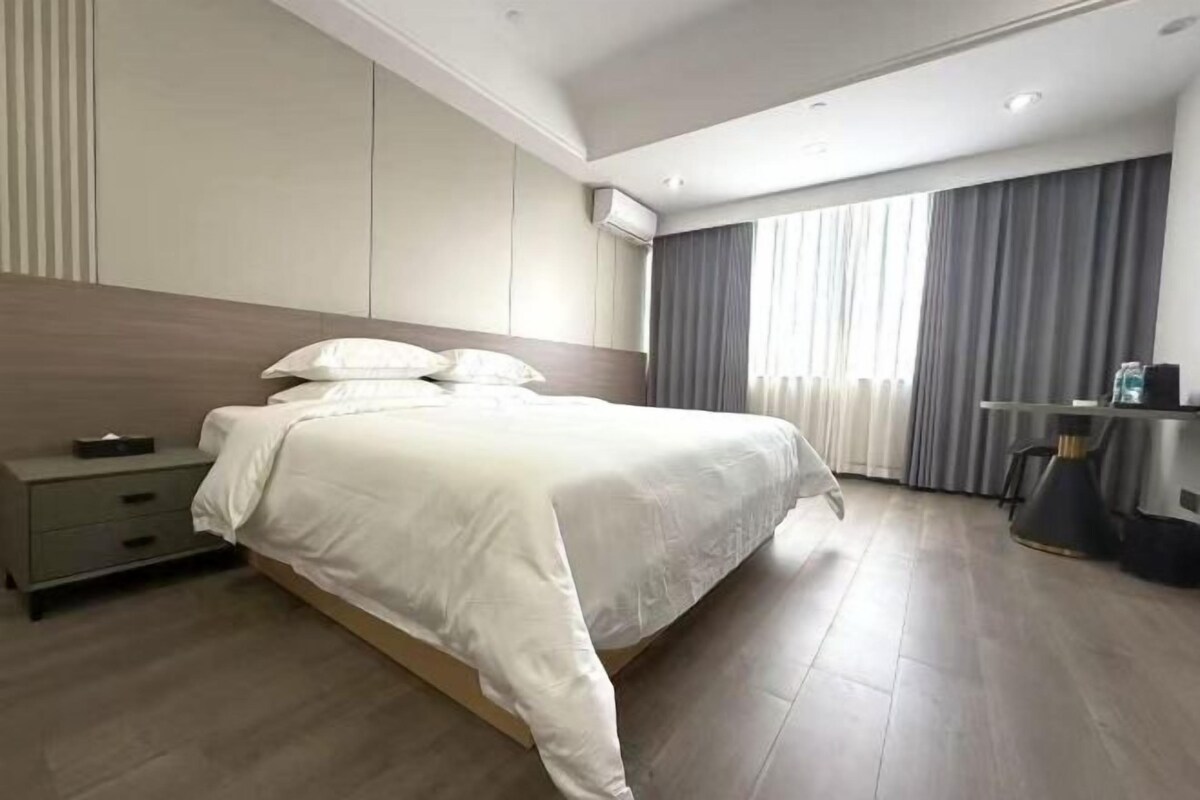 A modern hotel room with a large bed, a bedside table, a desk, and a window with curtains.