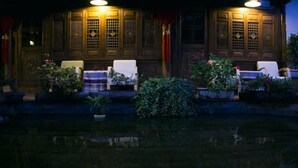 A traditional building with wooden doors and windows, outdoor seating with cushions, and a water feature in the foreground.