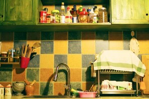 A kitchen with a tiled backsplash, a stainless steel sink, and various kitchen items on shelves and countertops. - Mrs. Sha's Living Room Inn (Jianchuan)