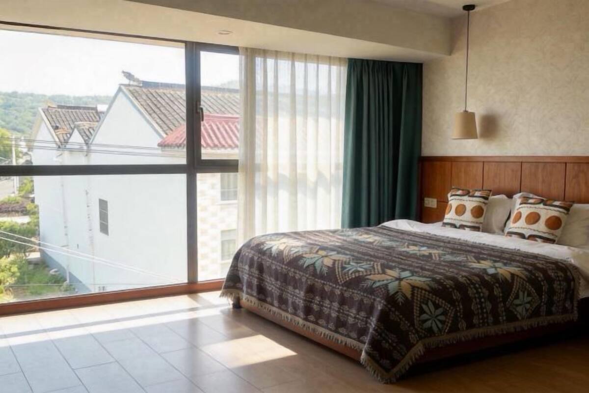 Quarto com uma janela grande, cama com roupa de cama estampada e vista para edifícios.