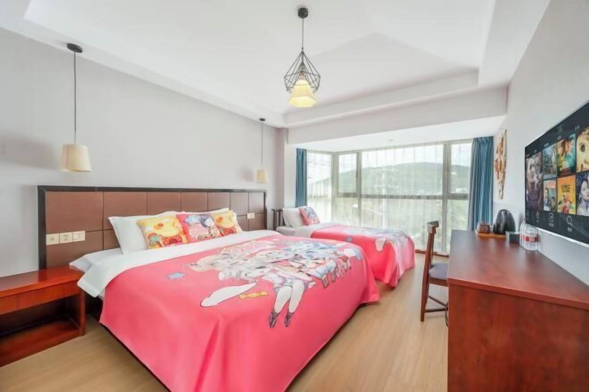 Quarto de hotel com duas camas, uma escrivaninha, uma janela grande e um lustre.