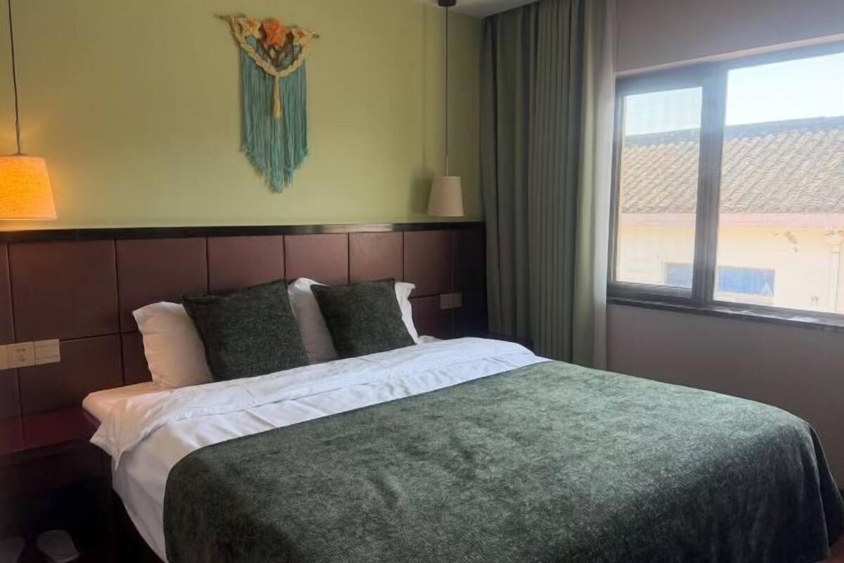 Quarto de hotel com cama, decoração em macramé fixada na parede, janela com cortinas e uma luminária.