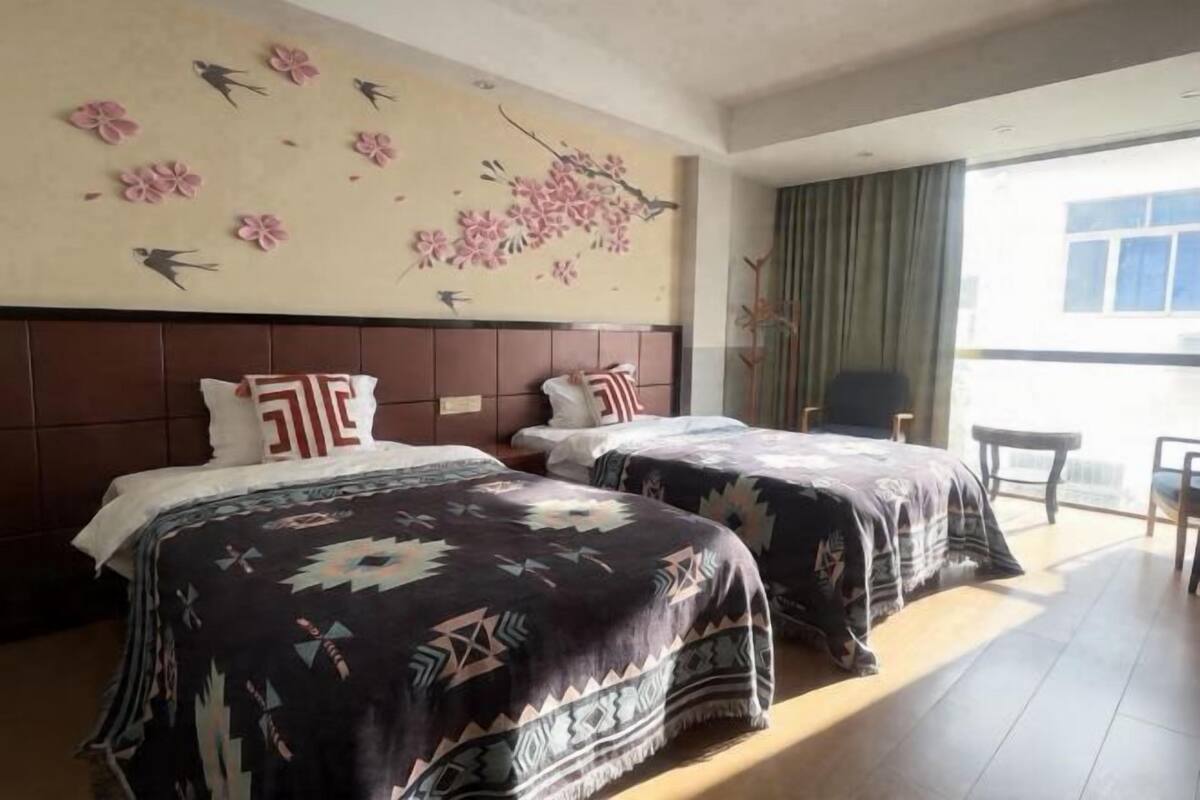 Quarto de hotel com duas camas, uma janela grande com cortinas, parede com decorações de flores e pássaros, e uma pequena mesa.