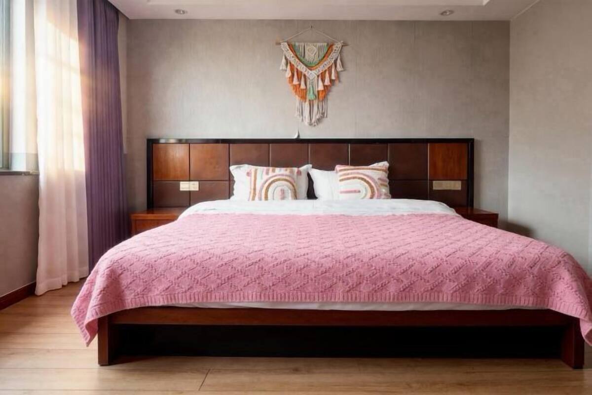 Quarto com cabeceira de madeira, colcha rosa e um painel de macramé na parede.