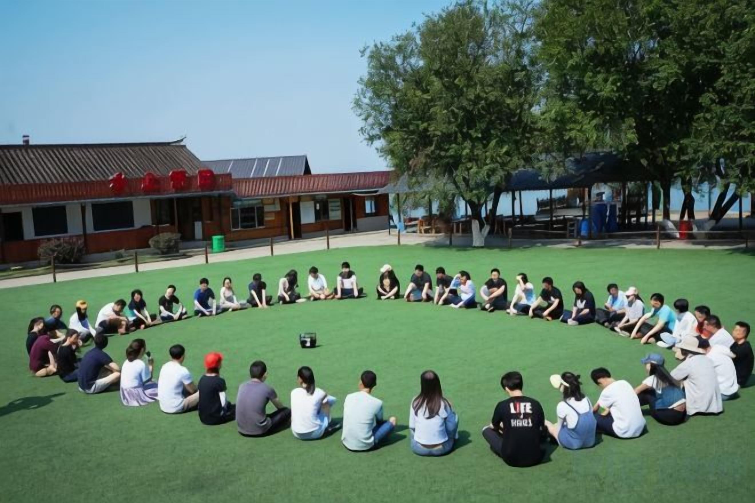 A group of people sitting in a circle on a grassy field in front of a building with a red roof.