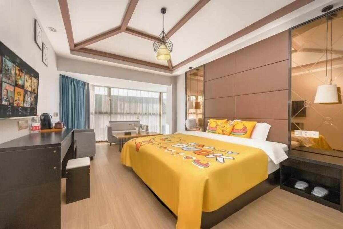 Quarto de hotel com cama, escrivaninha, cadeira, janela grande com cortinas e teto com vigas de madeira.