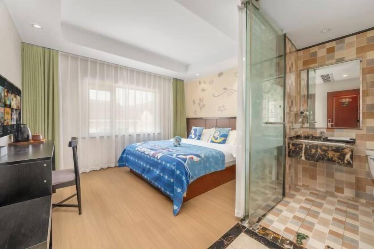 Quarto de hotel com cama, escrivaninha e banheiro com chuveiro.