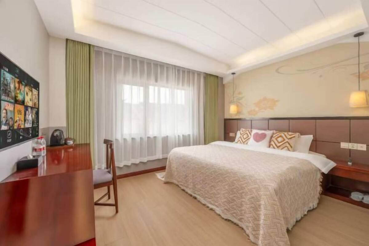 Quarto de hotel com cama, escrivaninha, cadeira, janela com cortinas e luminária na parede.