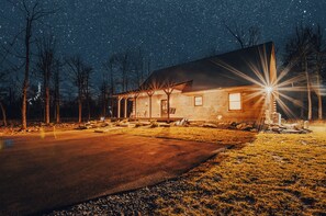 Une cabane en bois dotée d’un porche, d’une fenêtre et d’une porte, entourée d’arbres et surplombant un ciel étoilé.