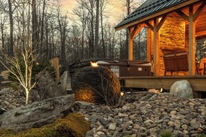 Une cheminée en pierre avec un feu qui crépite à l’intérieur d’un chalet en bois, lui-même entouré d’une forêt.