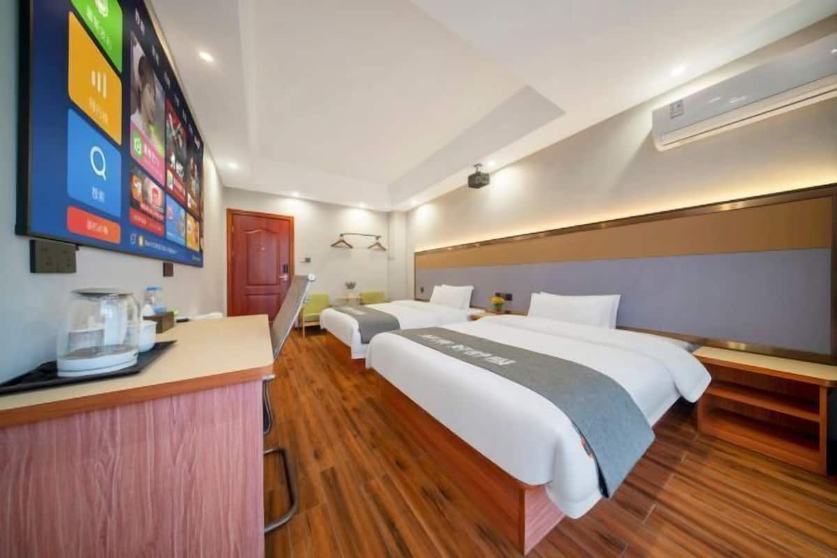 Habitación de hotel con piso de madera, dos camas, un escritorio con cafetera y un televisor de pared que muestra iconos de aplicaciones coloridos.