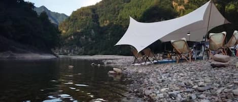 A tent set up on a rocky riverbank with chairs and a table, surrounded by a forested area.