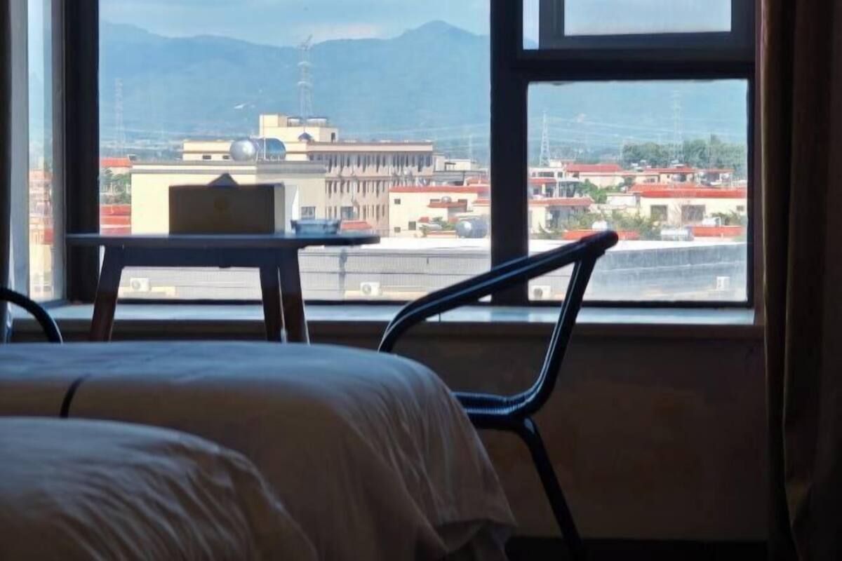 A hotel room with a bed, a desk, and a chair, with a view of buildings and mountains through a large window.