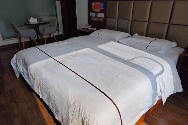 A large bed with white bedding and a brown headboard, a small round table with chairs, and a wall-mounted phone.
