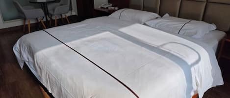 Una cama grande con ropa de cama blanca y un cabecero marrón, una mesita redonda con sillas, y un teléfono montado en la pared.