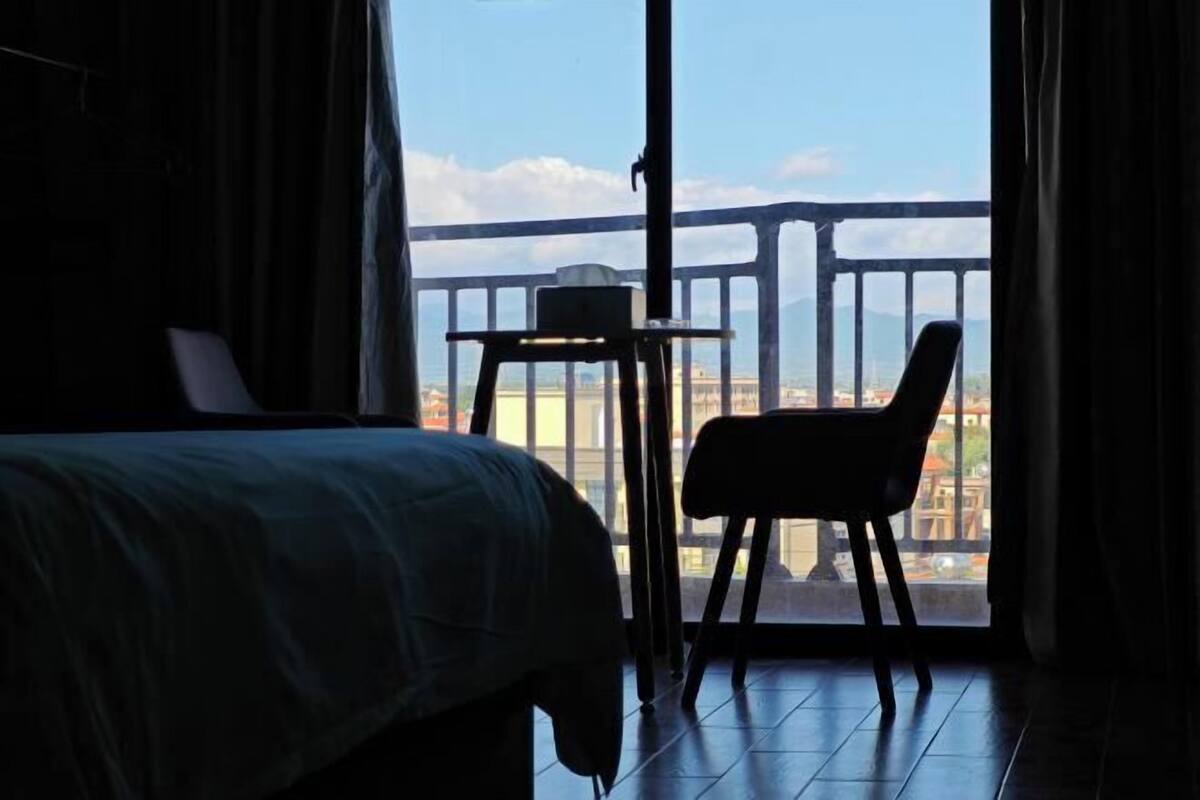 A room with a bed, a chair, a small table, and a balcony with a view.
