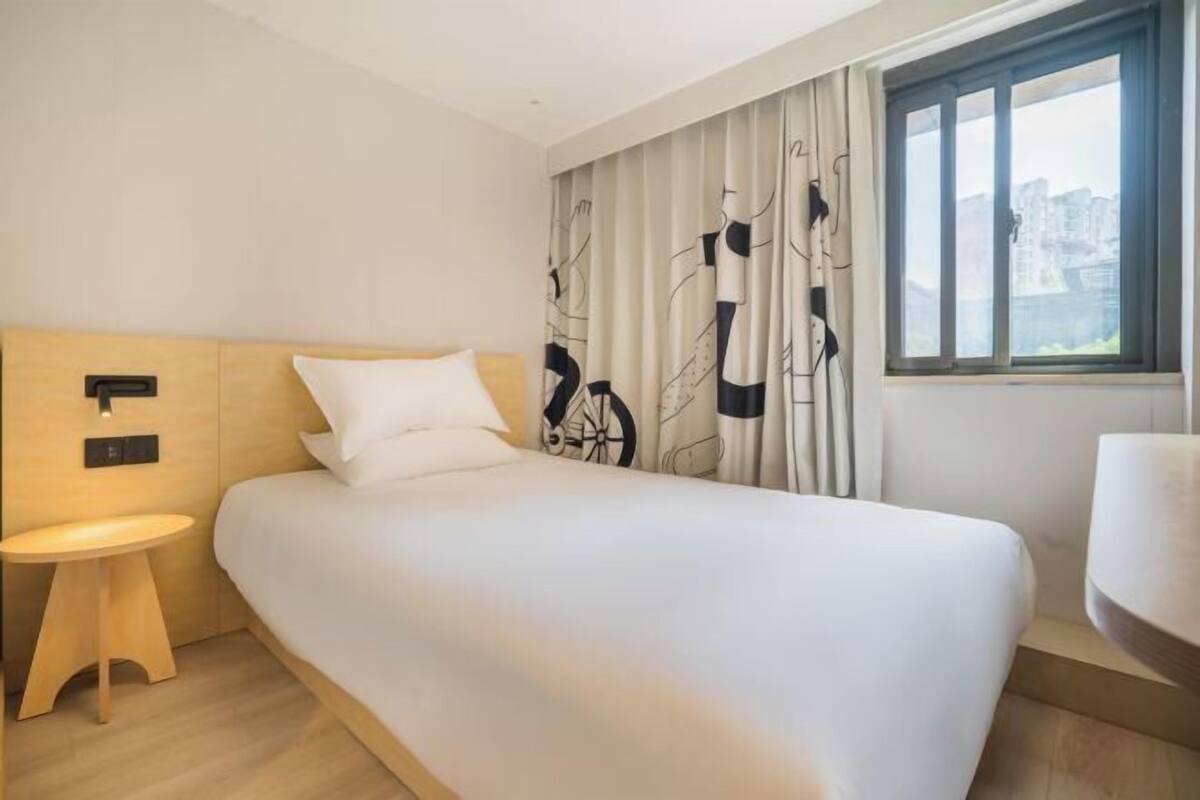 A modern hotel room with a large bed, a small bedside table, a wall-mounted control panel, and a window with a view of buildings.