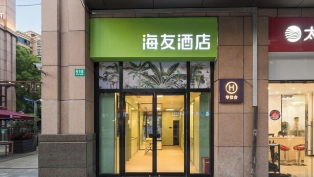 Entrada de hotel com placa verde, portas de vidro e interior visível com área de estar.