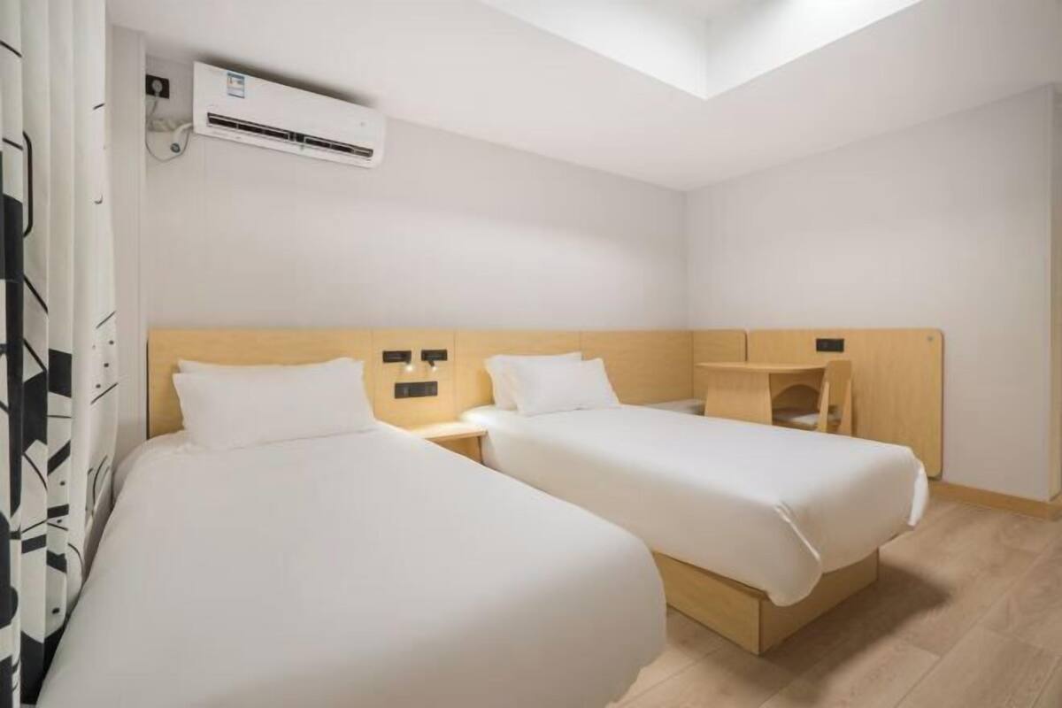 A hotel room with two single beds, a wooden headboard, a small desk, and an air conditioning unit.