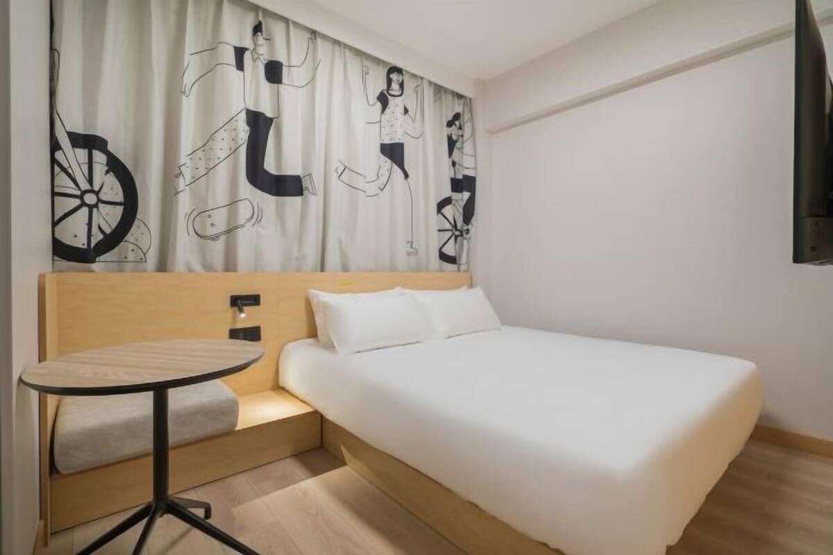 A hotel room with a bed, a small round table, and a wall with a curtain featuring abstract designs.