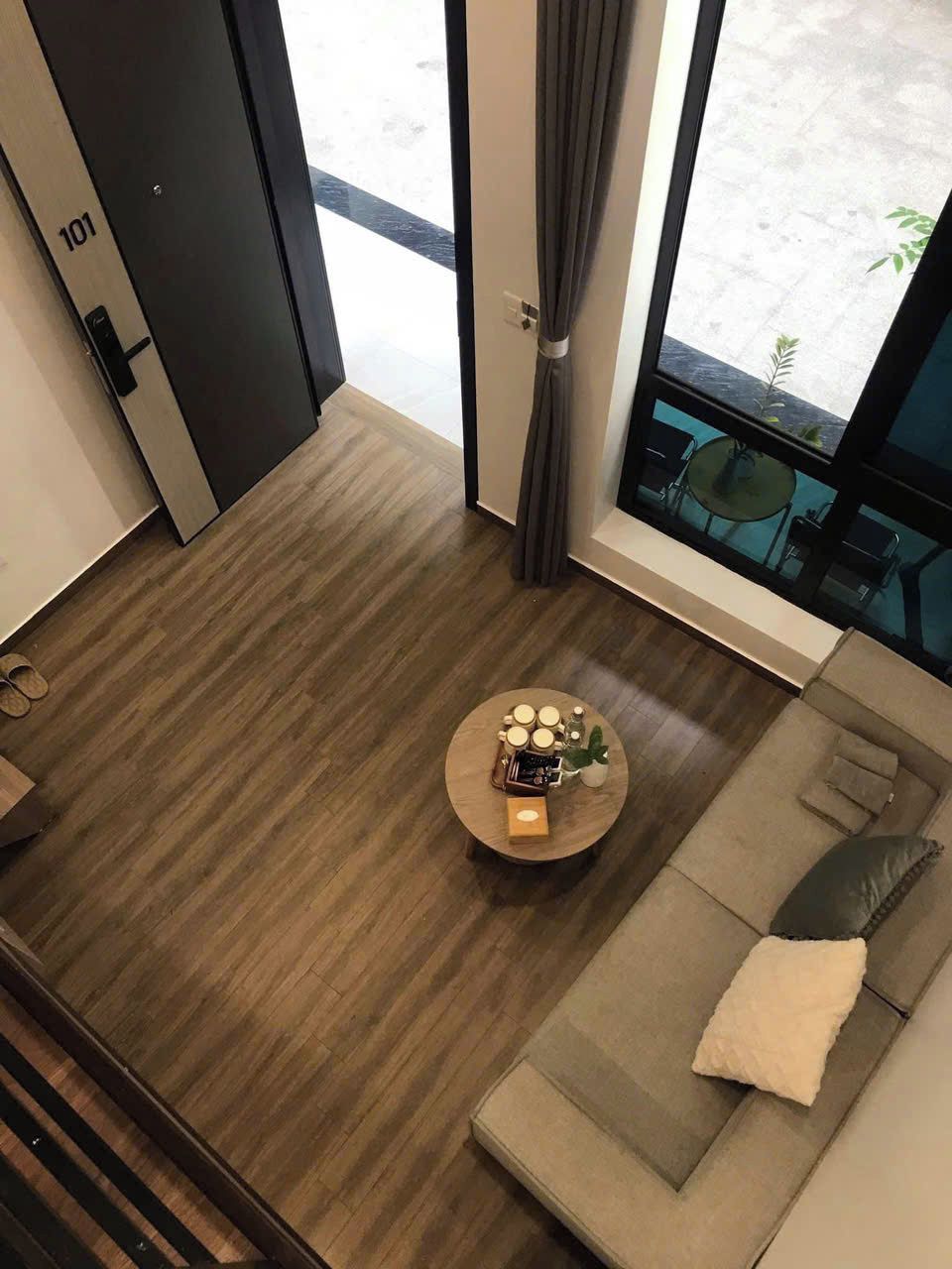 Une pièce moderne avec un plancher en bois, une table basse ronde sur laquelle sont disposés divers objets, un canapé avec des coussins et une vue sur l’extérieur grâce à une grande fenêtre.