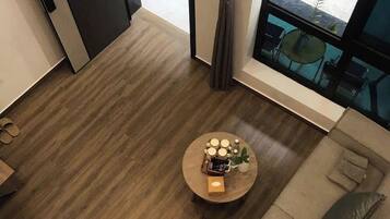Une pièce moderne avec un plancher en bois, une table basse ronde sur laquelle sont disposés divers objets, un canapé avec des coussins et une vue sur l’extérieur grâce à une grande fenêtre.