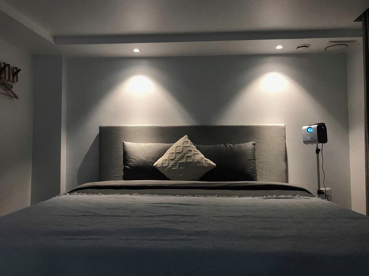 A modern bedroom with a large bed, two bedside lamps, and a ceiling-mounted projector.
