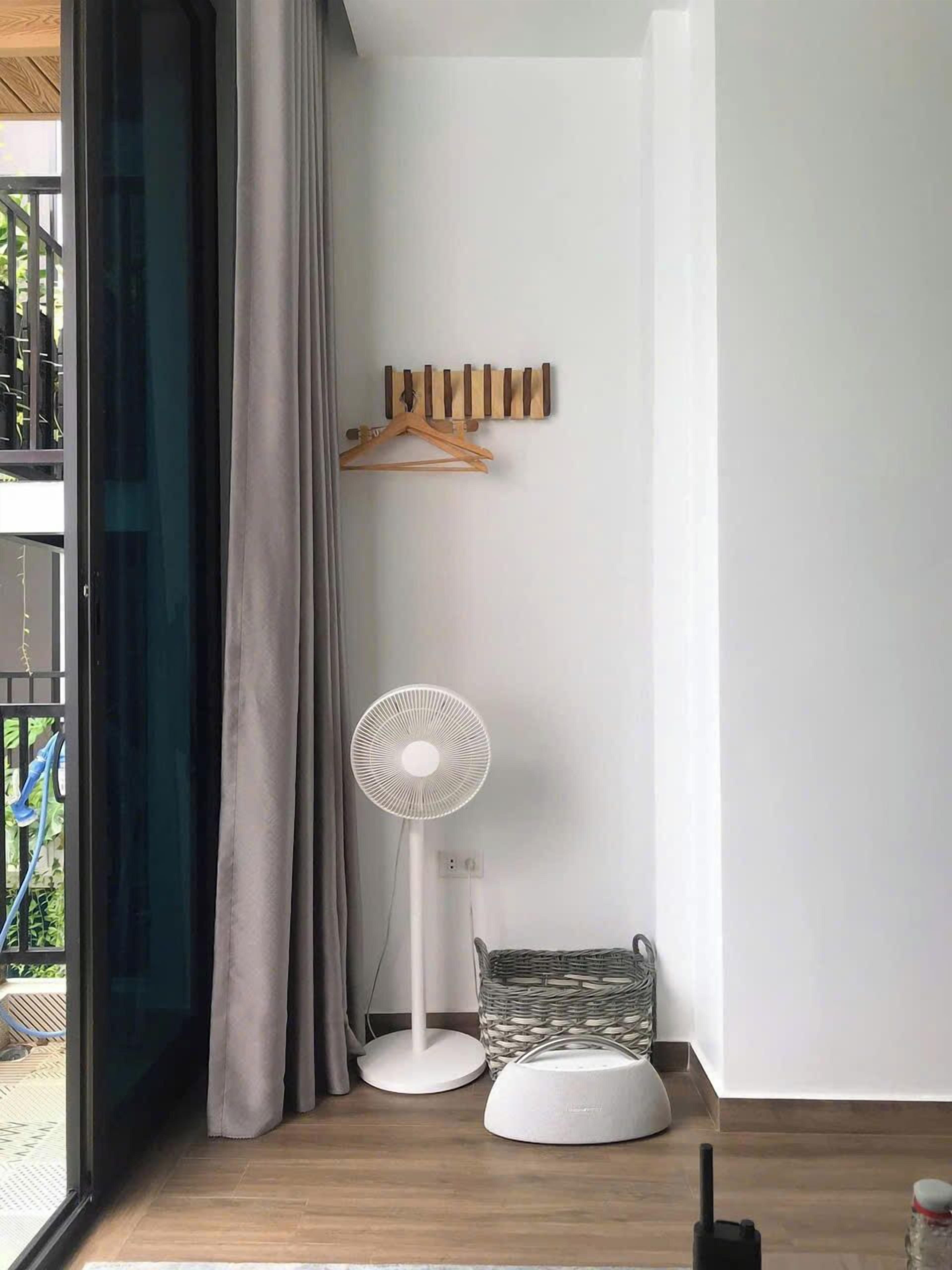 Ein Zimmer mit großem Fenster, einem Standventilator, einer weißen Lampe und einem Korbwägelchen auf dem Boden.
