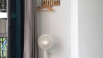 Ein Zimmer mit großem Fenster, einem Standventilator, einer weißen Lampe und einem Korbwägelchen auf dem Boden.