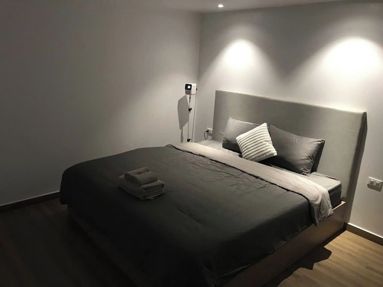 Una cama bien hecha con una colcha oscura, una toalla doblada y un cabecero con una lámpara.