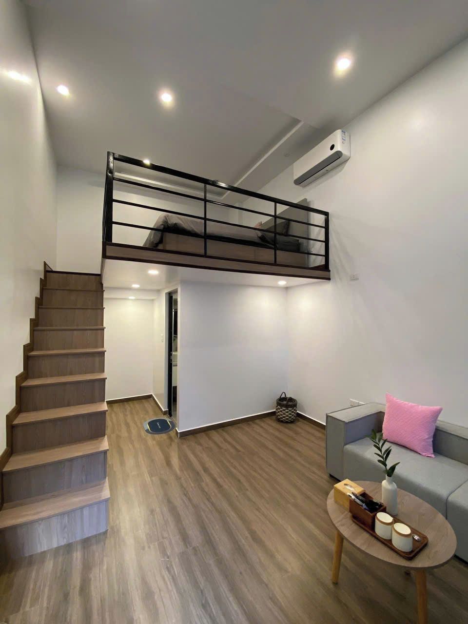 Un espacio habitable moderno con escalera de madera, zona superior con barandilla negra, sofá gris y una pequeña mesa redonda con objetos decorativos.