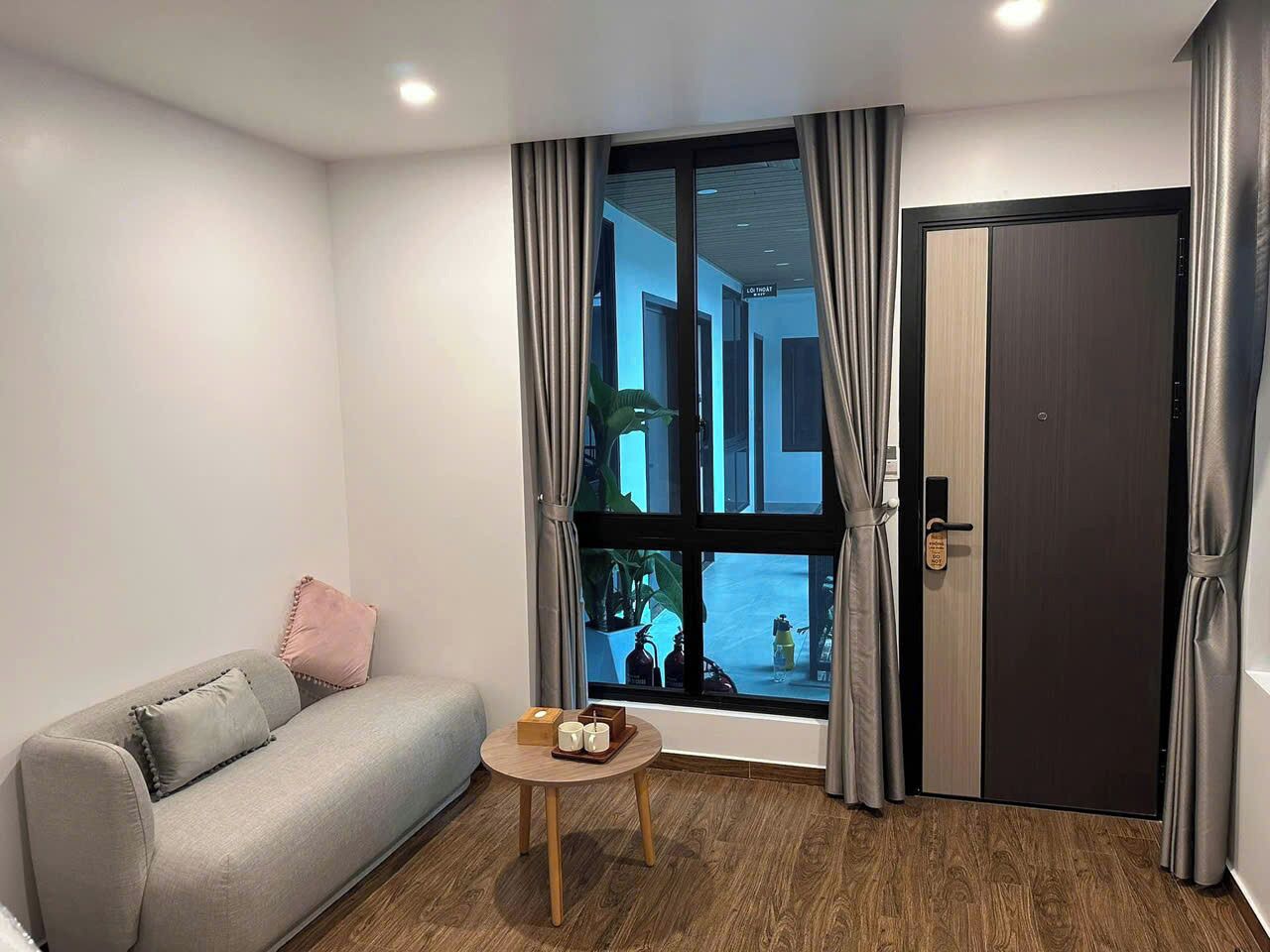 Una habitación moderna con un sofá gris, una pequeña mesa de madera con una bandeja y un ventanal con cortinas.