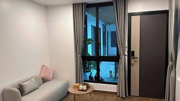 Una habitación moderna con un sofá gris, una pequeña mesa de madera con una bandeja y un ventanal con cortinas.