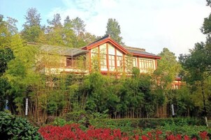 Un edificio de madera tradicional con ventanas amplias y un balcón cubierto, rodeado de exuberante vegetación y follaje rojo.