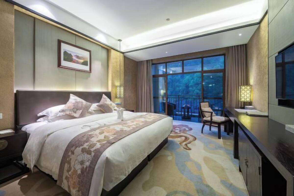 A hotel room with a large bed, a desk with a chair, a TV, and a balcony with a view.