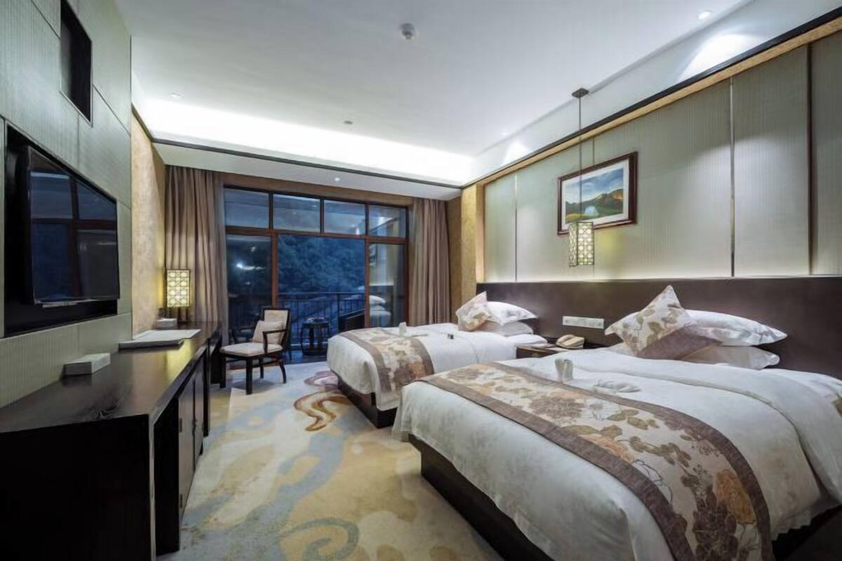 A hotel room with two beds, a desk with a TV, a chair, a balcony, and a painting on the wall.