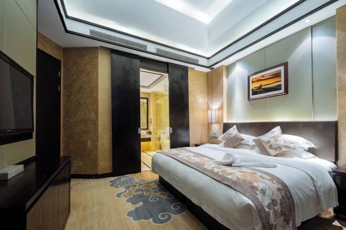A hotel room with a bed, a TV, a door, and a painting on the wall.