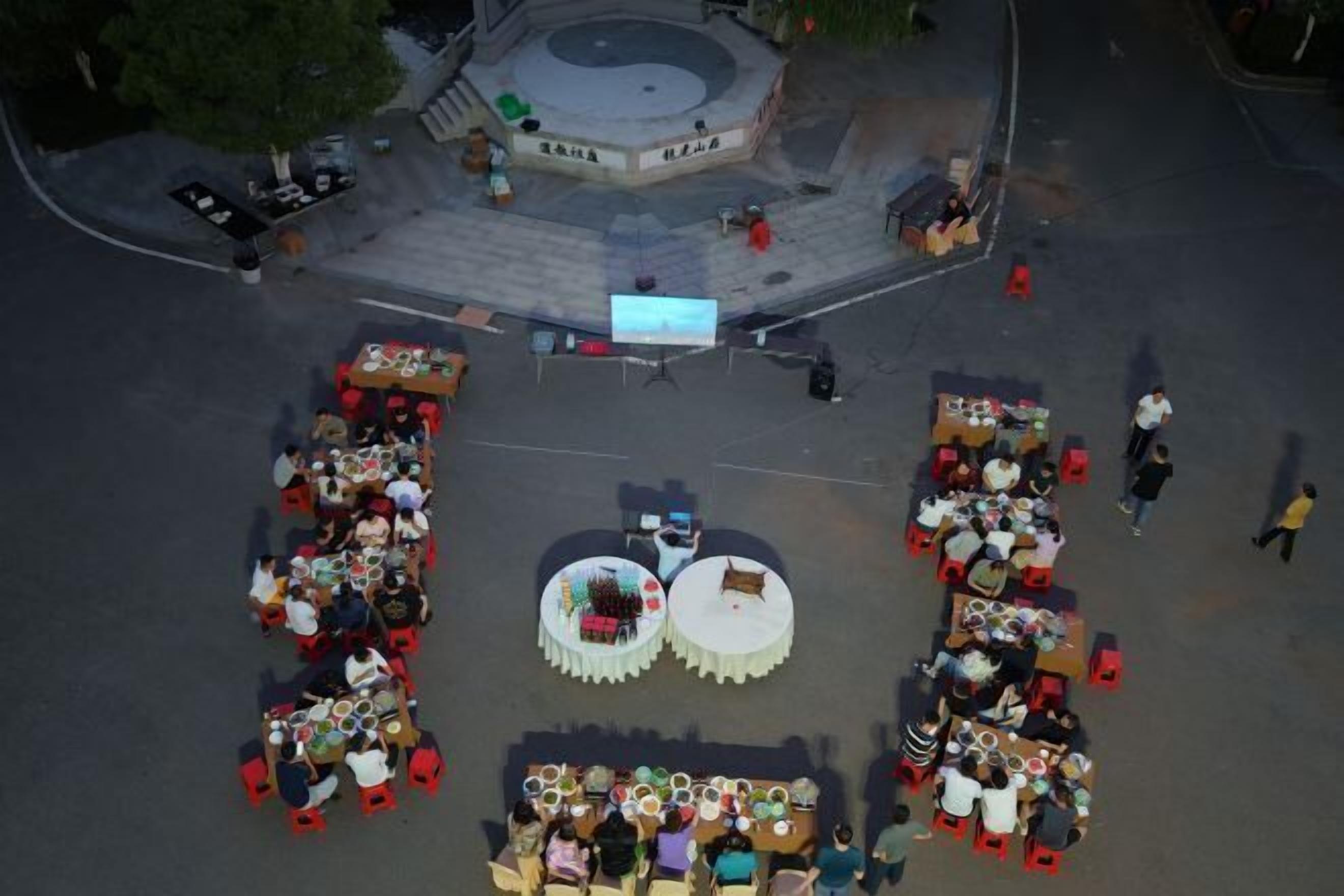 Un área de comedor al aire libre con varias mesas y sillas redondas, rodeada por una plaza pavimentada con una fuente central.