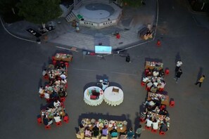 An outdoor dining area with multiple round tables and chairs, surrounded by a paved plaza with a central fountain.
