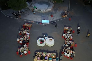 Un área de comedor al aire libre con varias mesas y sillas redondas, rodeada por una plaza pavimentada con una fuente central.