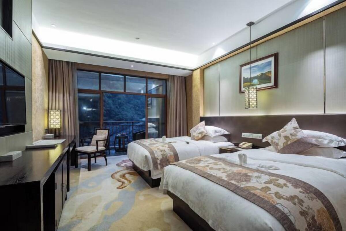 A hotel room with two beds, a desk, a chair, a TV, and a large window with a view.