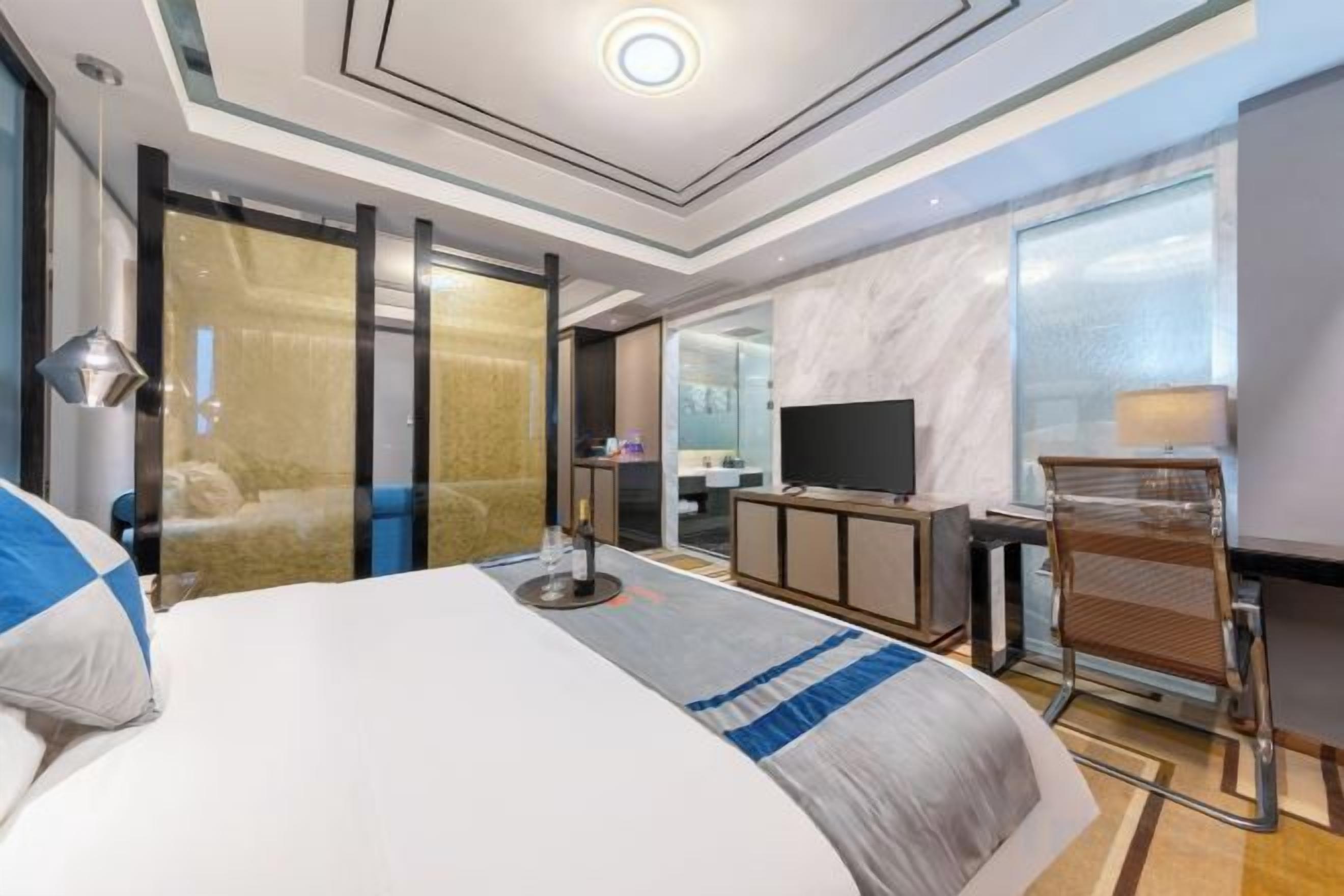 Ein modernes Hotelzimmer mit einem großen Bett, einem Schreibtisch, einem Fernseher und einem durch eine Glastür einsehbaren Badezimmer.
