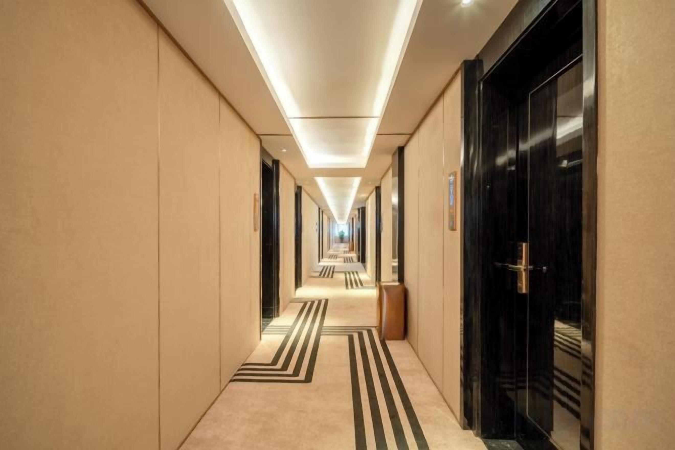 A hotel corridor with a long, straight layout, featuring a carpeted floor with a striped pattern, and doors on both sides leading to rooms.