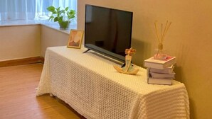 A room with a television on a stand, a small table with a decorative item, a framed picture, and a potted plant by the window. - Luzui Hotel (Shenzhen)