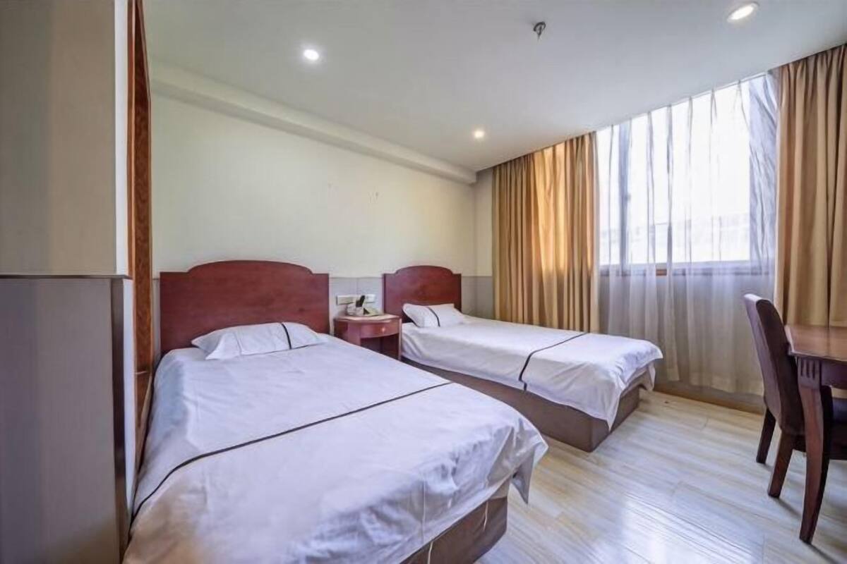 A hotel room with two beds, a wooden desk, and a large window with curtains.