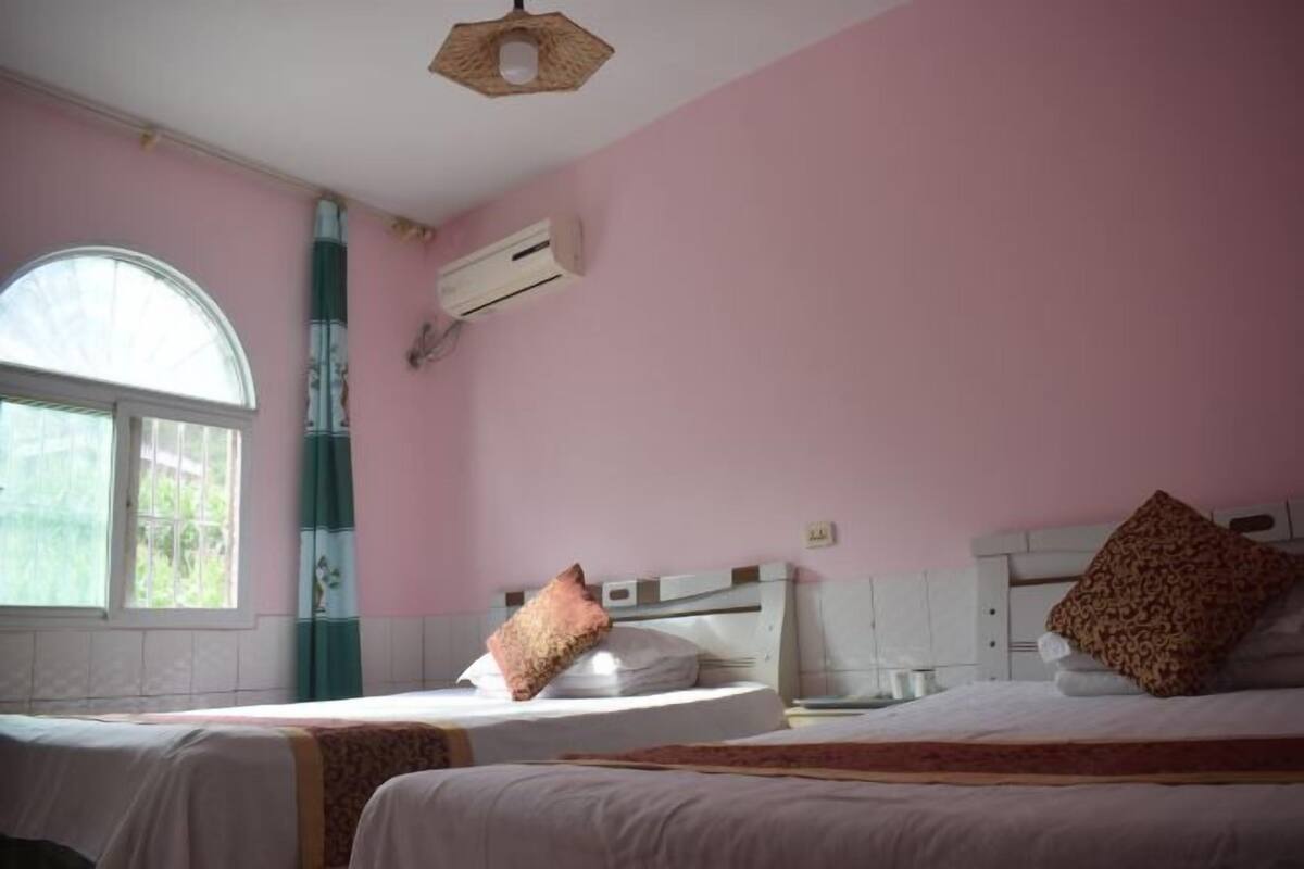 A bedroom with two beds, a window with a view, an air conditioning unit, and a wooden ceiling light fixture.
