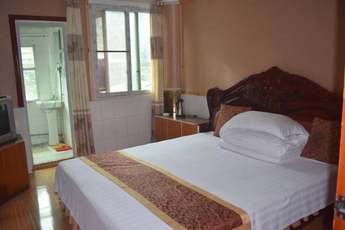 A bedroom with a bed, a wooden headboard, a window, a television, and a bathroom visible through an open door.